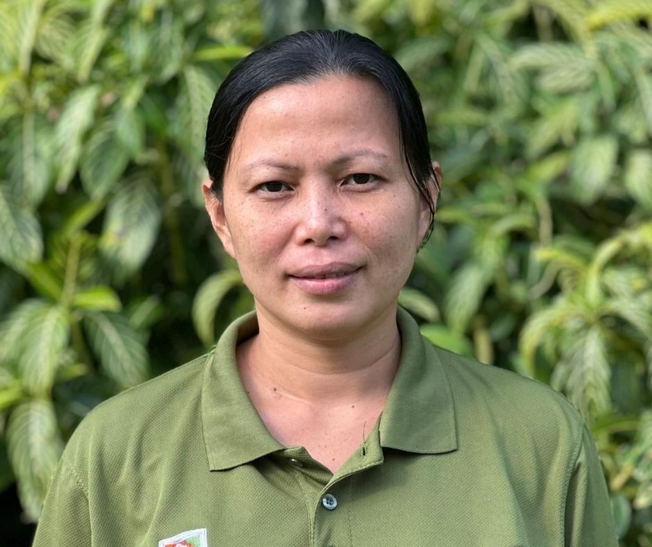 A woman standing and smiling for the camera with a green leafy bush in the background.