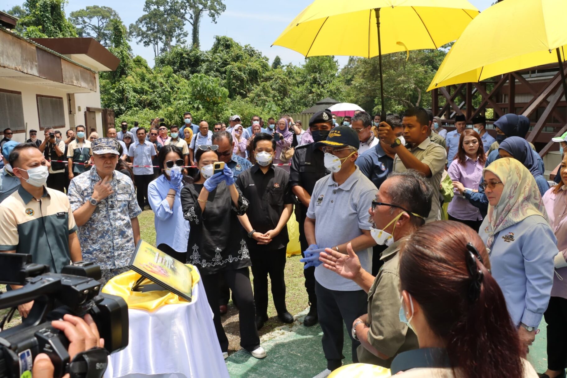 The king of Malaysia surrounded by staff, press, and government officials whilst he looks at a poster.