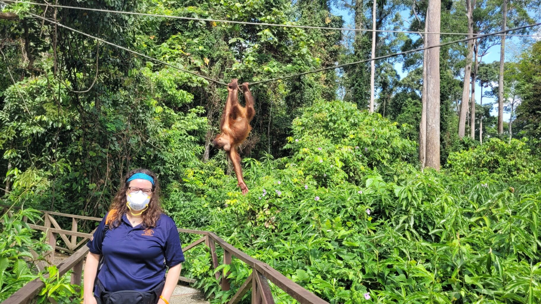 Manger for the charity, Adrienne stands for a photo with an orangutan on ropes climbing in the background.