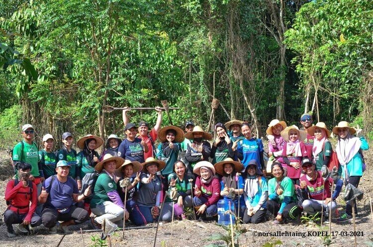 A large group of people who work to restore rainforests posing for a photo in front of tree planting fields.